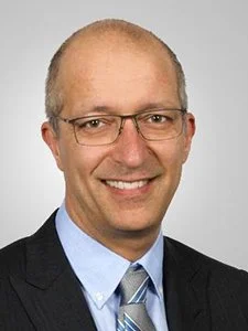 Portrait of a smiling man with glasses, wearing a dark suit and tie against a light background.
