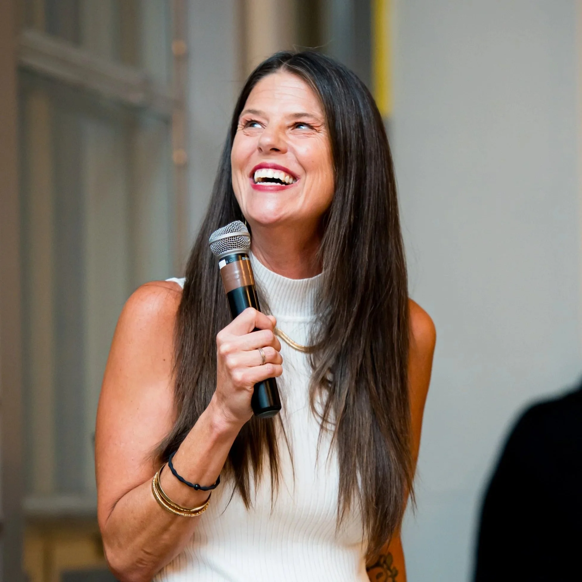 A woman with long brown hair, smiling, holding a microphone, wearing a sleeveless white top and gold jewelry, standing indoors in front of a blurred background.