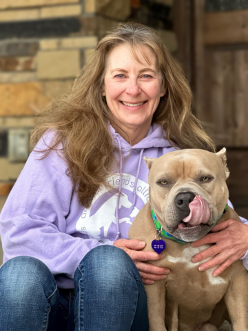 A smiling woman with wavy blonde hair wearing a light purple hoodie sitting with her arm around a large tan and white dog with a green collar on a porch.