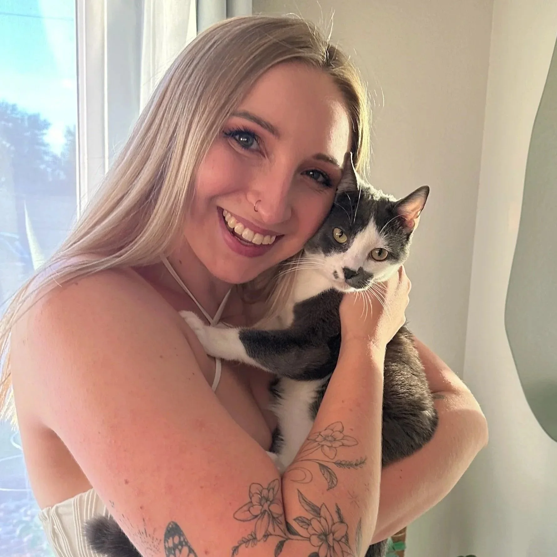 A woman with blonde hair smiling and hugging a gray and white cat indoors.