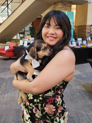 Young woman smiling and holding a small brown and black puppy with a white ribbon on its collar inside a building.