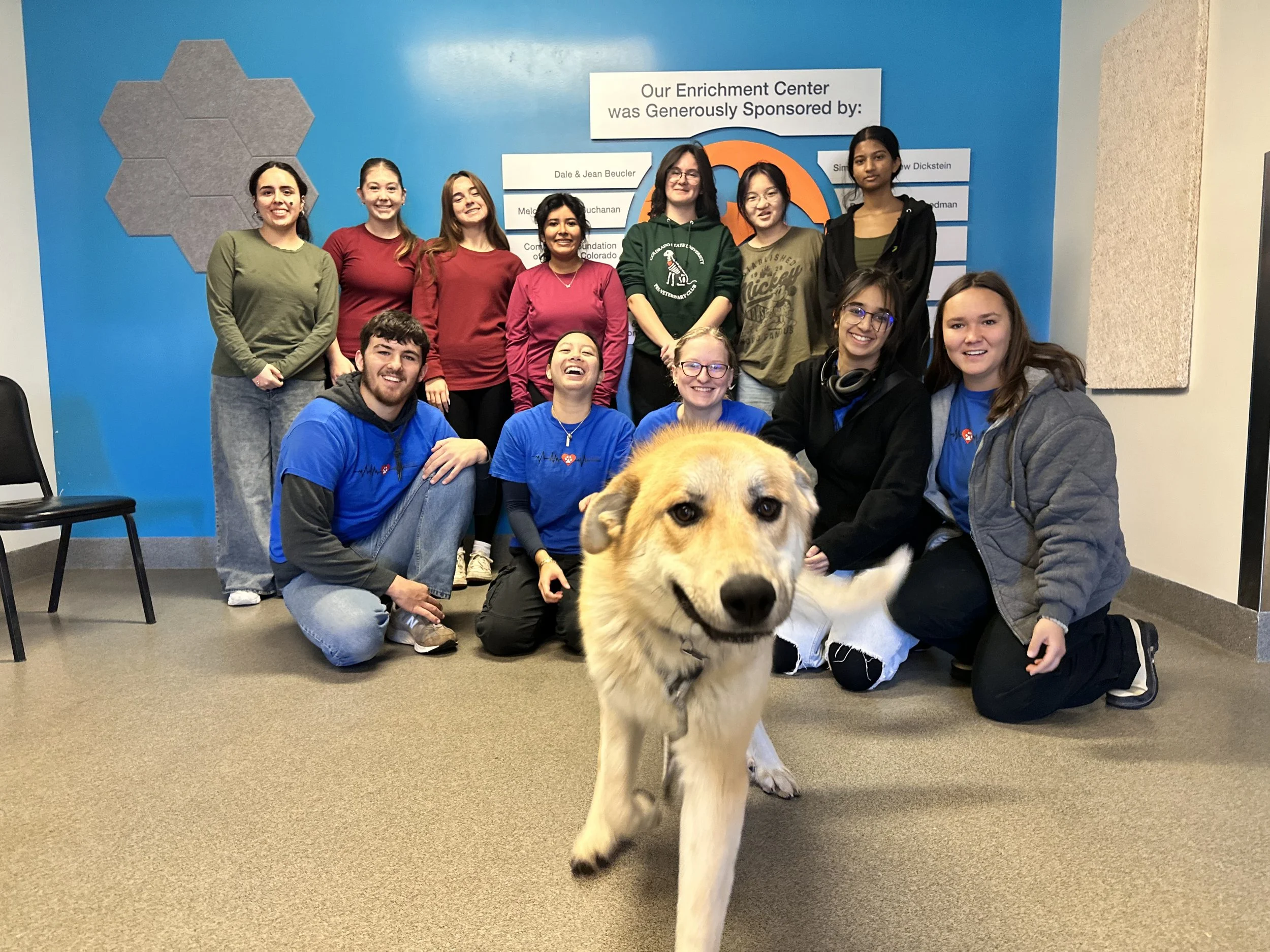 Group of people smiling around a service dog in a room with a blue wall and a sign that reads "Our Enrichment Center was generously sponsored by."