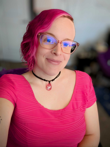 Young woman with pink hair, wearing glasses, a black choker with a pink and white pendant, and a pink top.