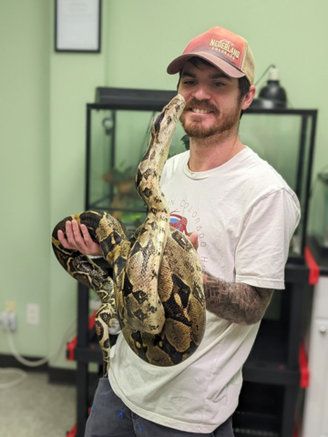 A man with a beard wearing a baseball cap and white t-shirt holding a large snake indoors.