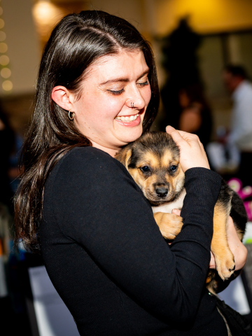 A woman with dark hair smiling and holding a small puppy indoor with blurred background.