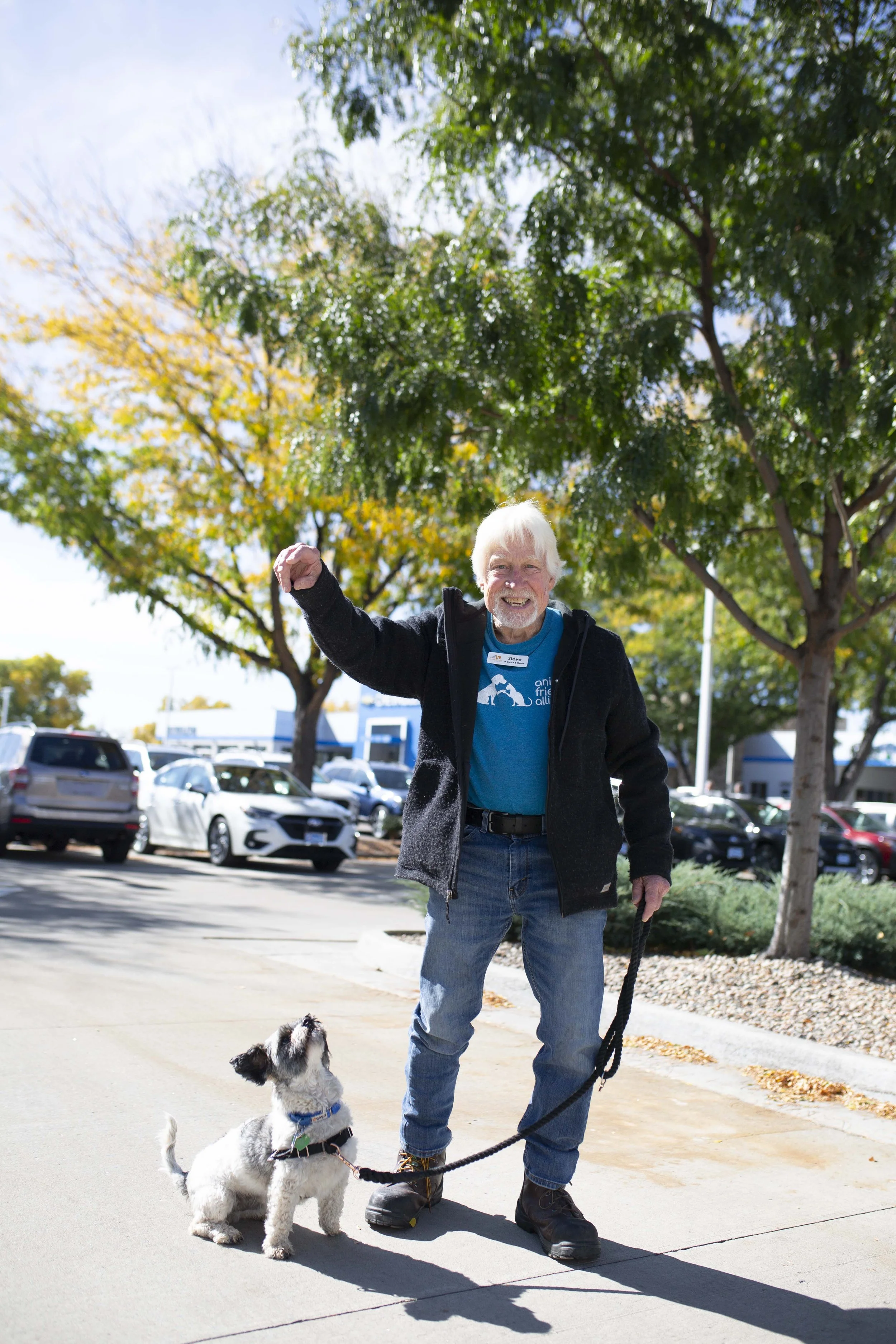 An elderly man with white hair and a beard, wearing a black jacket and blue t-shirt, is walking a small dog in a parking lot under green trees on a sunny day. The dog appears to be a white and black terrier mix.