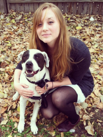 A young woman crouching next to a dog with a black and white coat, both sitting on fallen autumn leaves in a backyard.