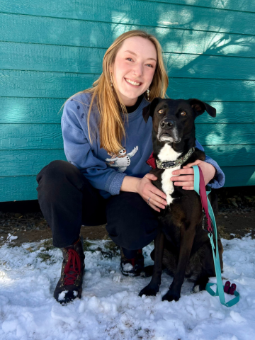 A young woman with long blonde hair smiling and kneeling on snow next to a black and white dog in front of a teal wooden wall.