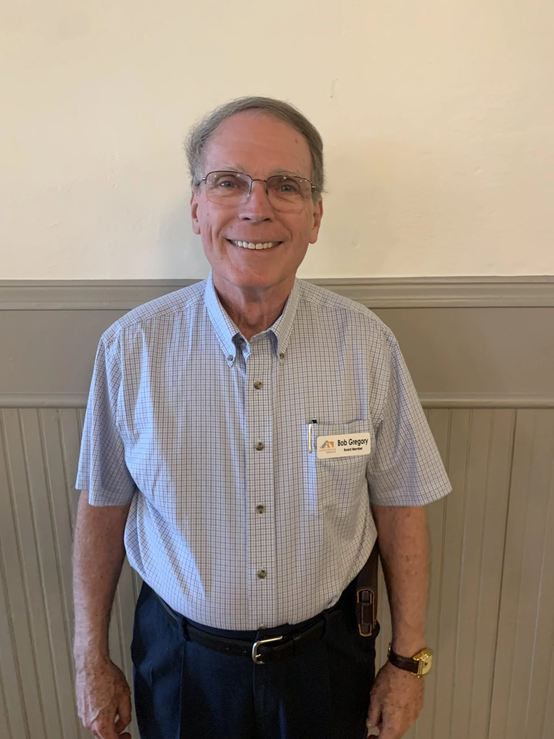A smiling elderly man wearing glasses, a light blue checkered shirt, dark pants, and a watch, standing indoors against a paneled wall. He has a name tag that reads 'Bob Gregory'.