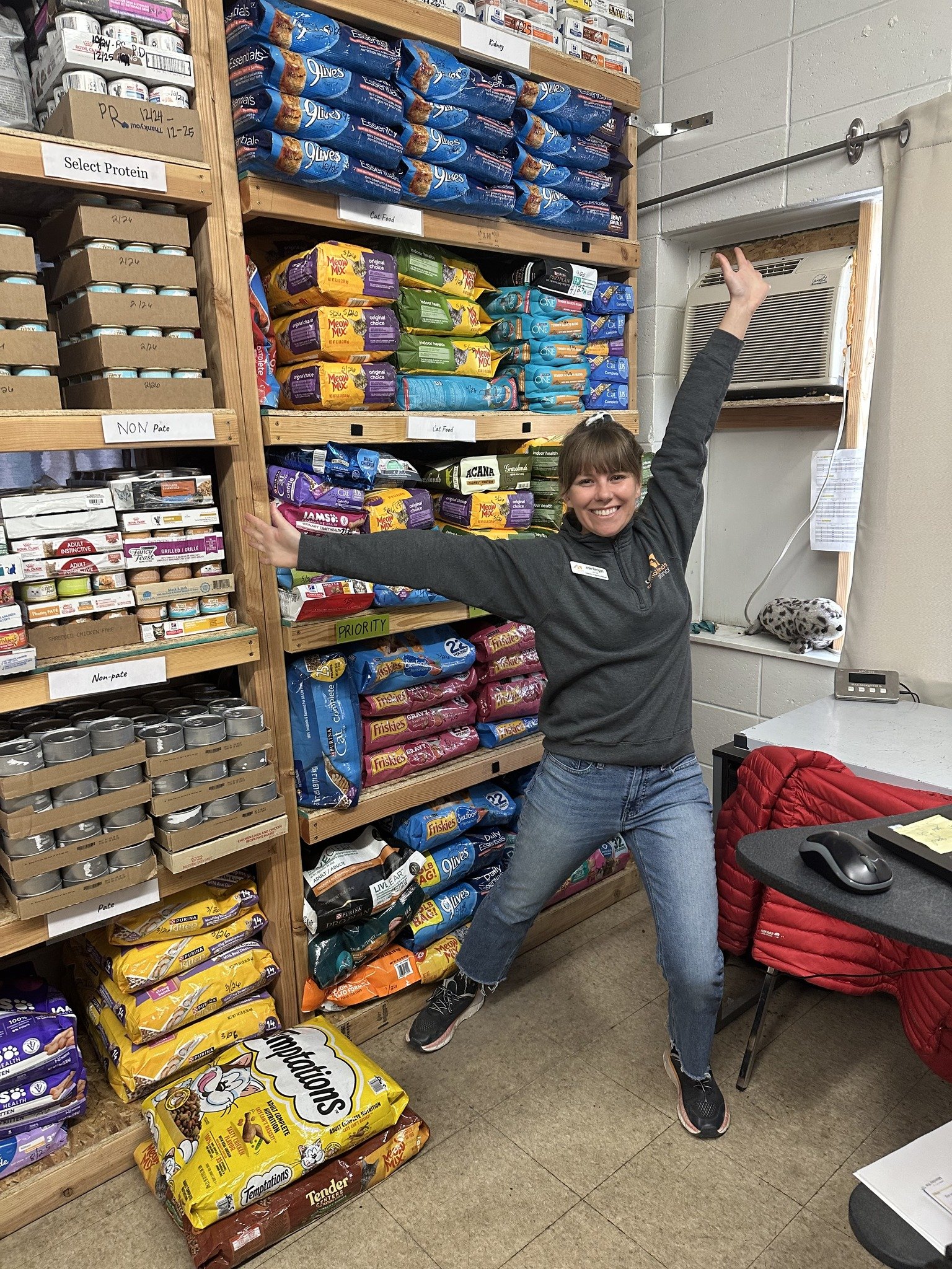 A woman standing in a pet store or animal care aisle, cheerfully posing with arms outstretched in front of shelves stocked with bags of pet food and canned pet food, with a window air conditioning unit and a small amount of clutter nearby.