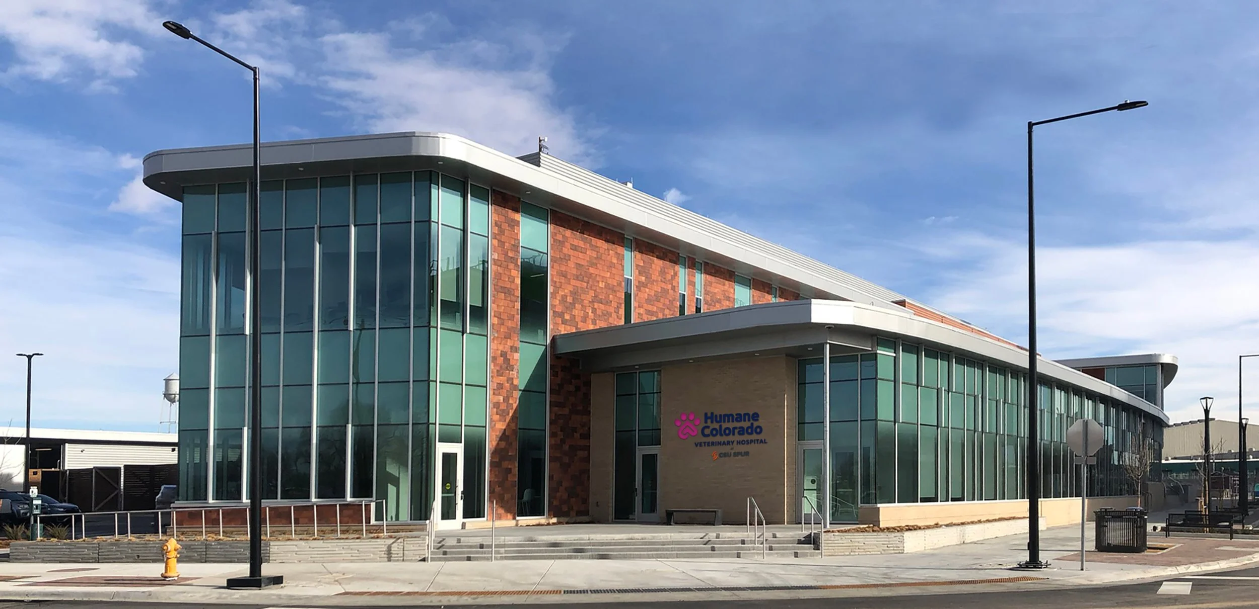 Modern veterinary hospital building with large glass windows, brick accents, and a sign that reads "Humane Colorado Veterinary Hospital" in purple and orange, situated in a sunny urban area with lamp posts and a few parked cars.