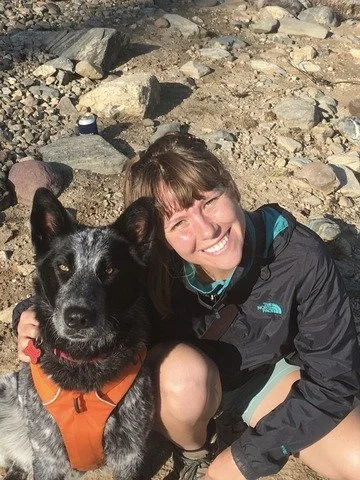 A smiling woman in outdoor clothing crouching next to a large black and gray dog with a collar and harness, on rocky terrain.