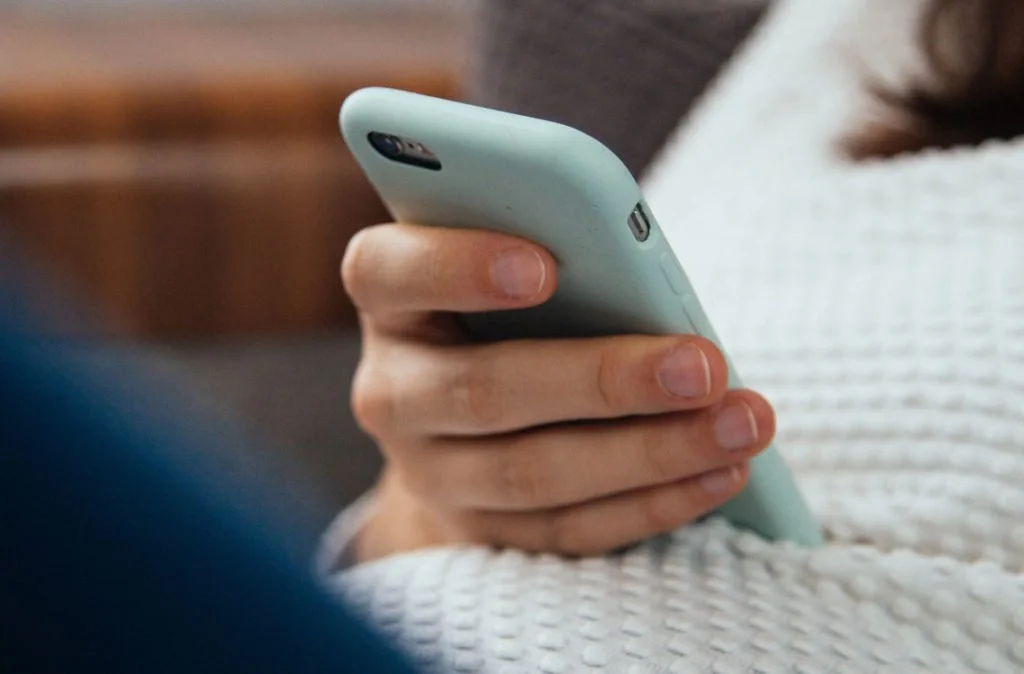 Close-up of a person holding a light blue smartphone in their hand, wearing a white knit sweater.