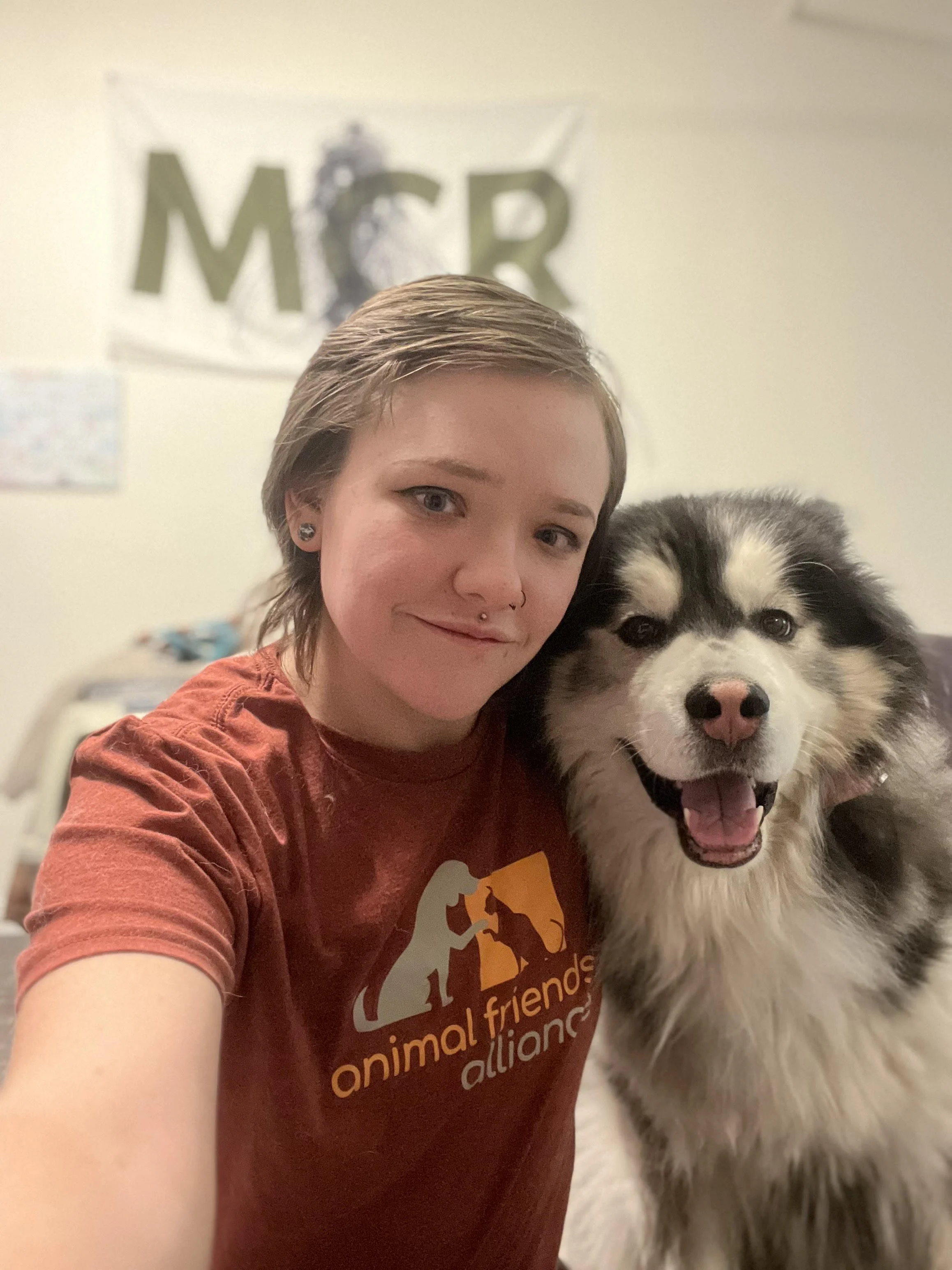 A young woman with short hair and a septum piercing smiling next to a happy Alaskan Malamute dog inside a room with a banner that reads 'MCR' in the background. The woman is wearing a red t-shirt with the logo of the Animal Friends Alliance.