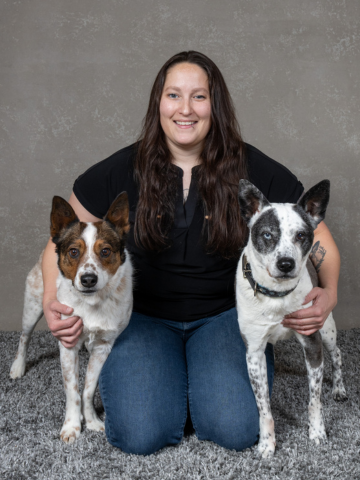 A woman with long brown hair kneeling on a gray carpet, smiling, holding two dogs, one on each side, in front of a gray textured wall.