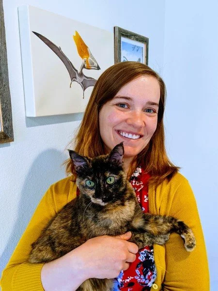 A smiling woman holding a grey and black cat indoors, with a dinosaur painting on the wall behind her.