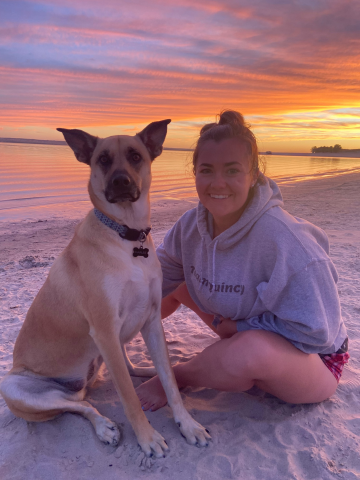A young woman and a dog sitting on a sandy beach at sunset, with colorful sky and water in the background.