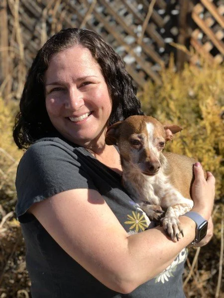 Woman smiling and holding a small brown and white dog outdoors with bushes in the background.