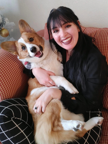 A woman with black hair and a big smile holding a happy corgi dog on her lap, sitting on a red checkered couch.