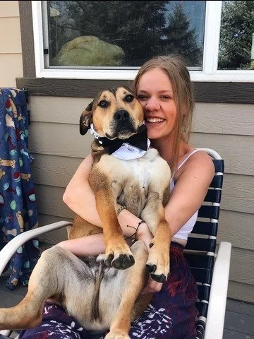Young woman smiling and hugging a large mixed-breed dog with a bow tie, sitting on a chair outside a house.
