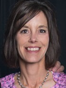 Close-up of a smiling woman with shoulder-length dark hair, wearing a purple patterned blouse and a pearl necklace.