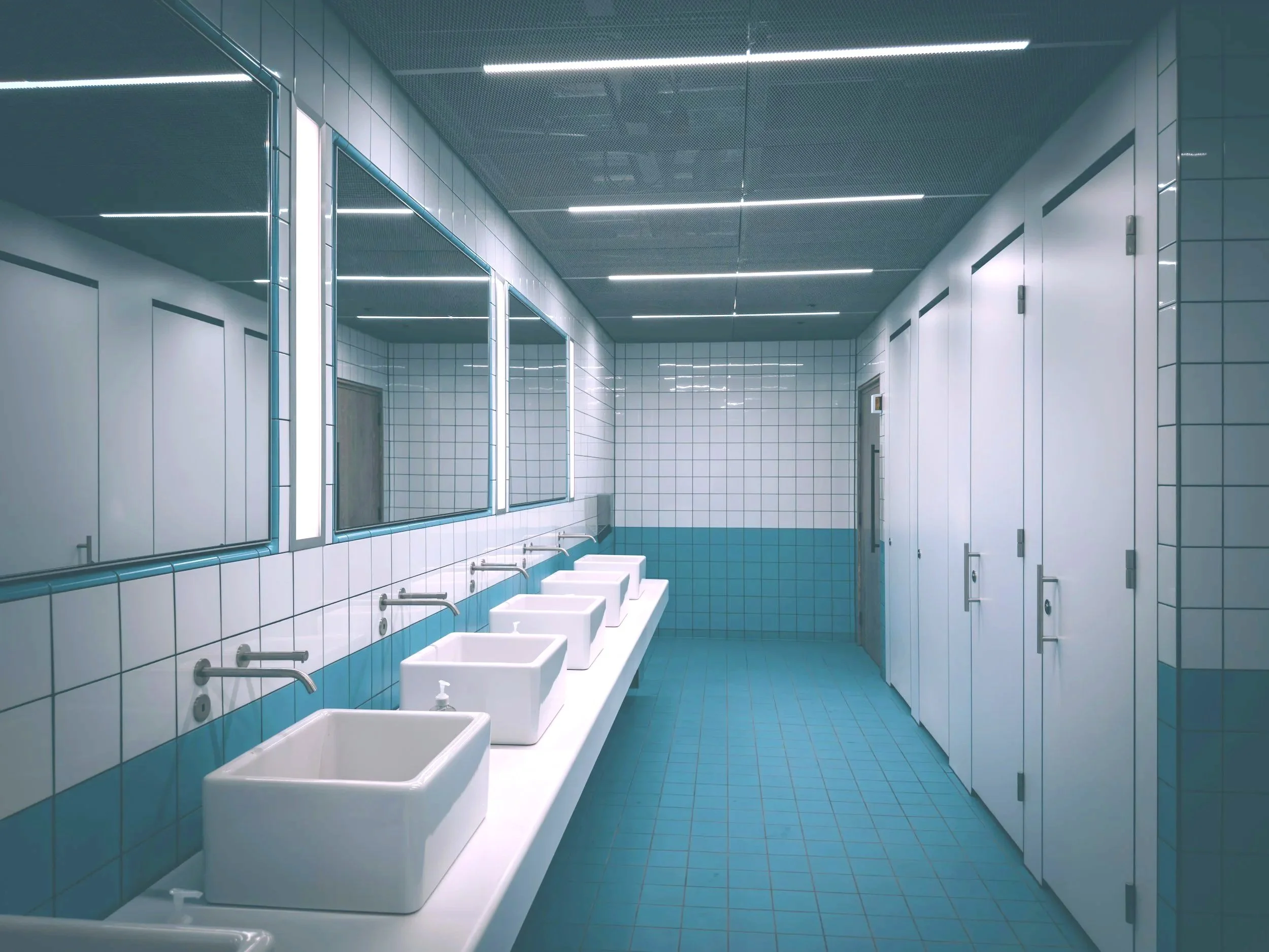 Public restroom with six white sinks along a blue and white tiled wall, large mirrors above the sinks, and white stall doors on the right side.