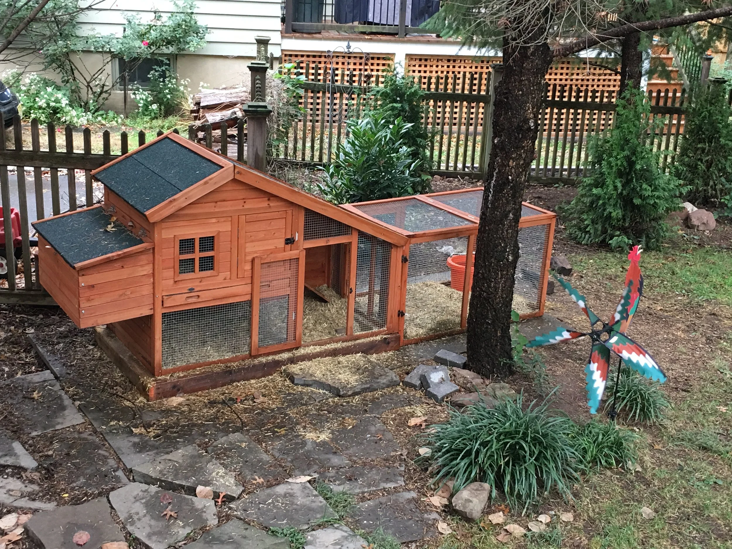 A wooden outdoor chicken coop with a fenced enclosure, a small window, and a water bucket. There is a colorful pinwheel and some plants nearby, and the area is surrounded by a wooden fence and trees.