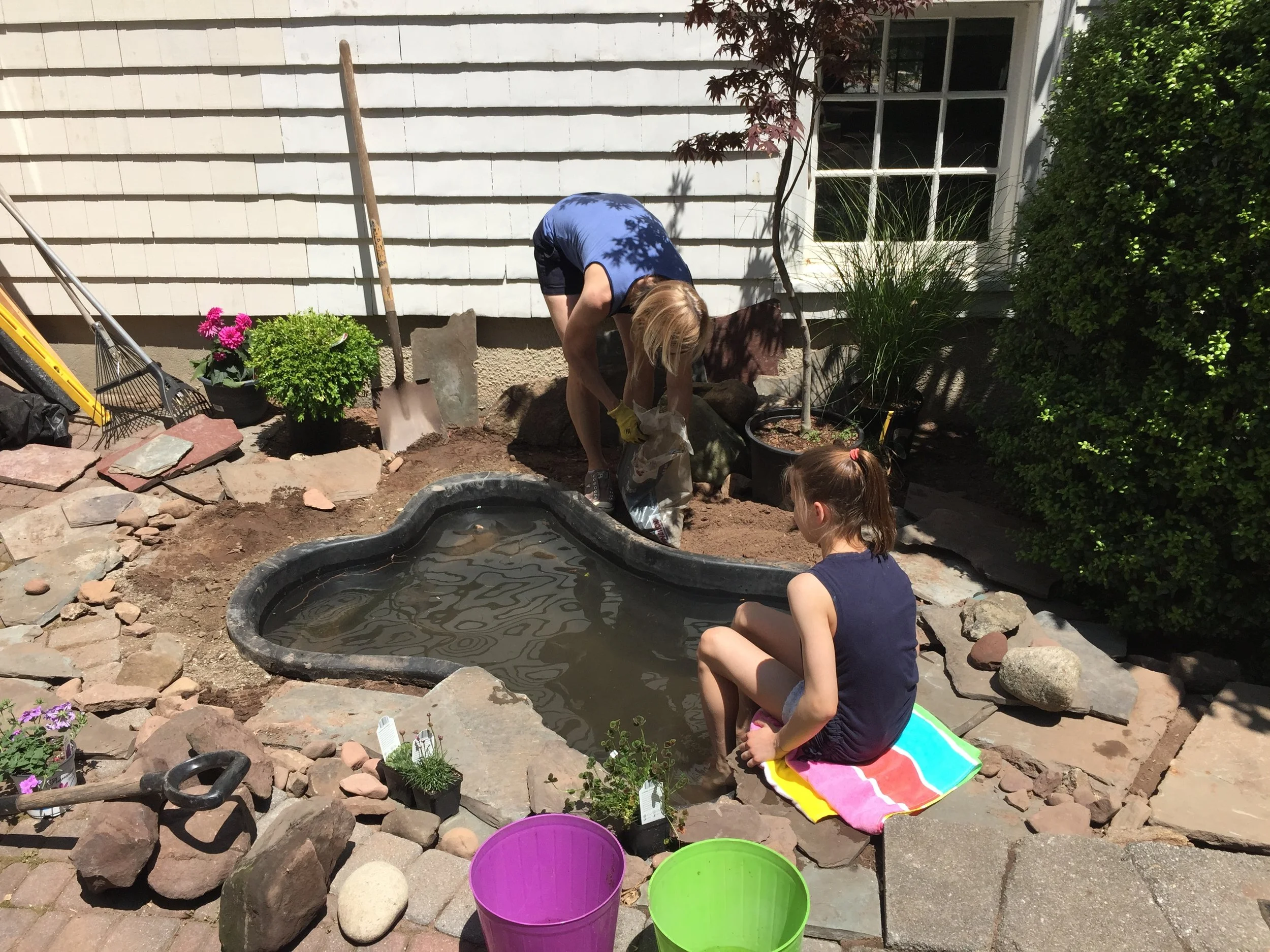A woman and a girl are working on a garden pond in a backyard. The woman is filling a bag, possibly with soil or gravel, while the girl is sitting on a colorful towel near the pond, observing. Gardening tools, potted plants, and buckets are visible nearby. The scene is sunlit with a white house and greenery in the background.
