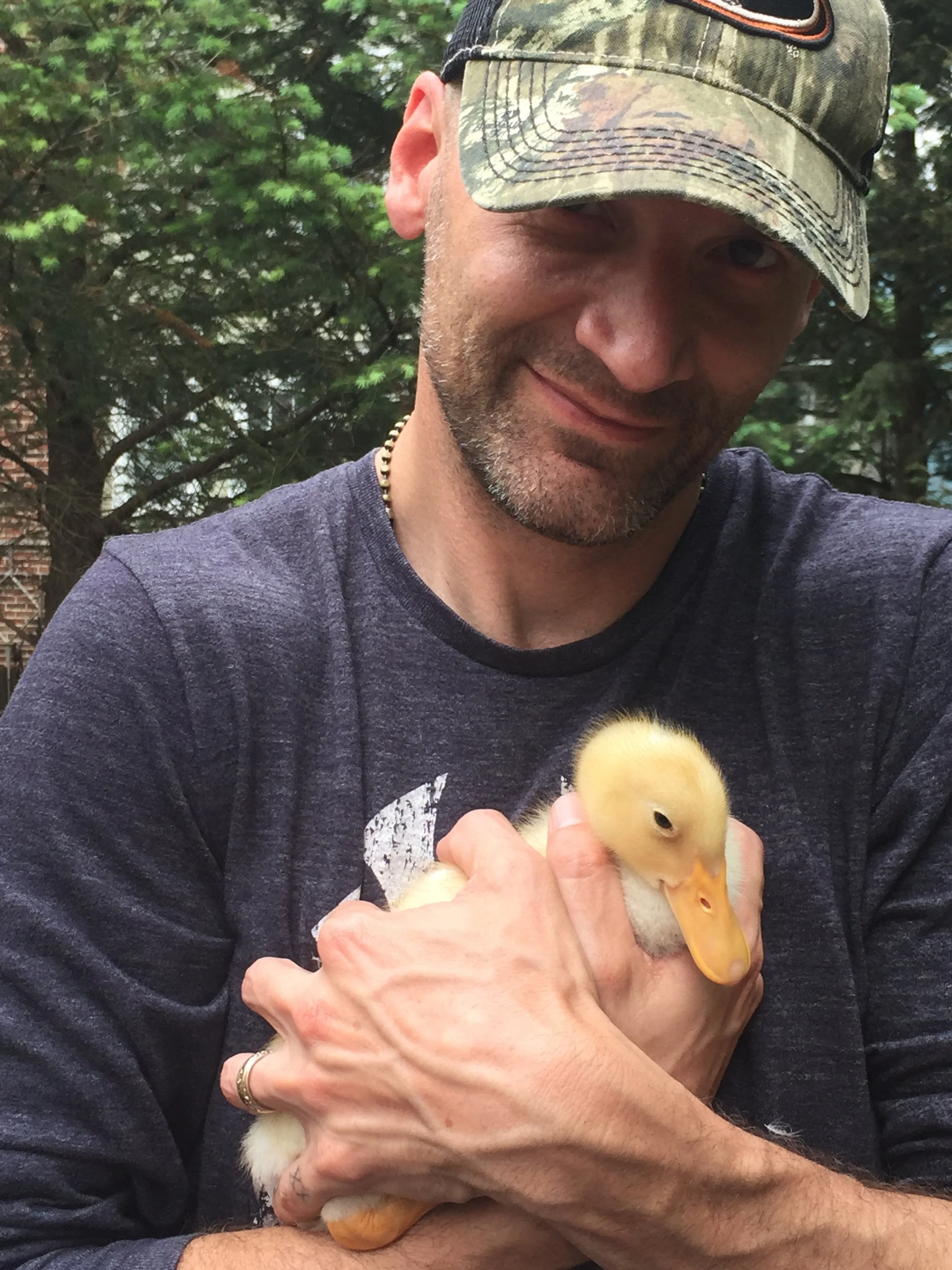 Man wearing a camo baseball cap and a dark gray shirt, holding a small yellow duckling, outdoors with trees in the background.