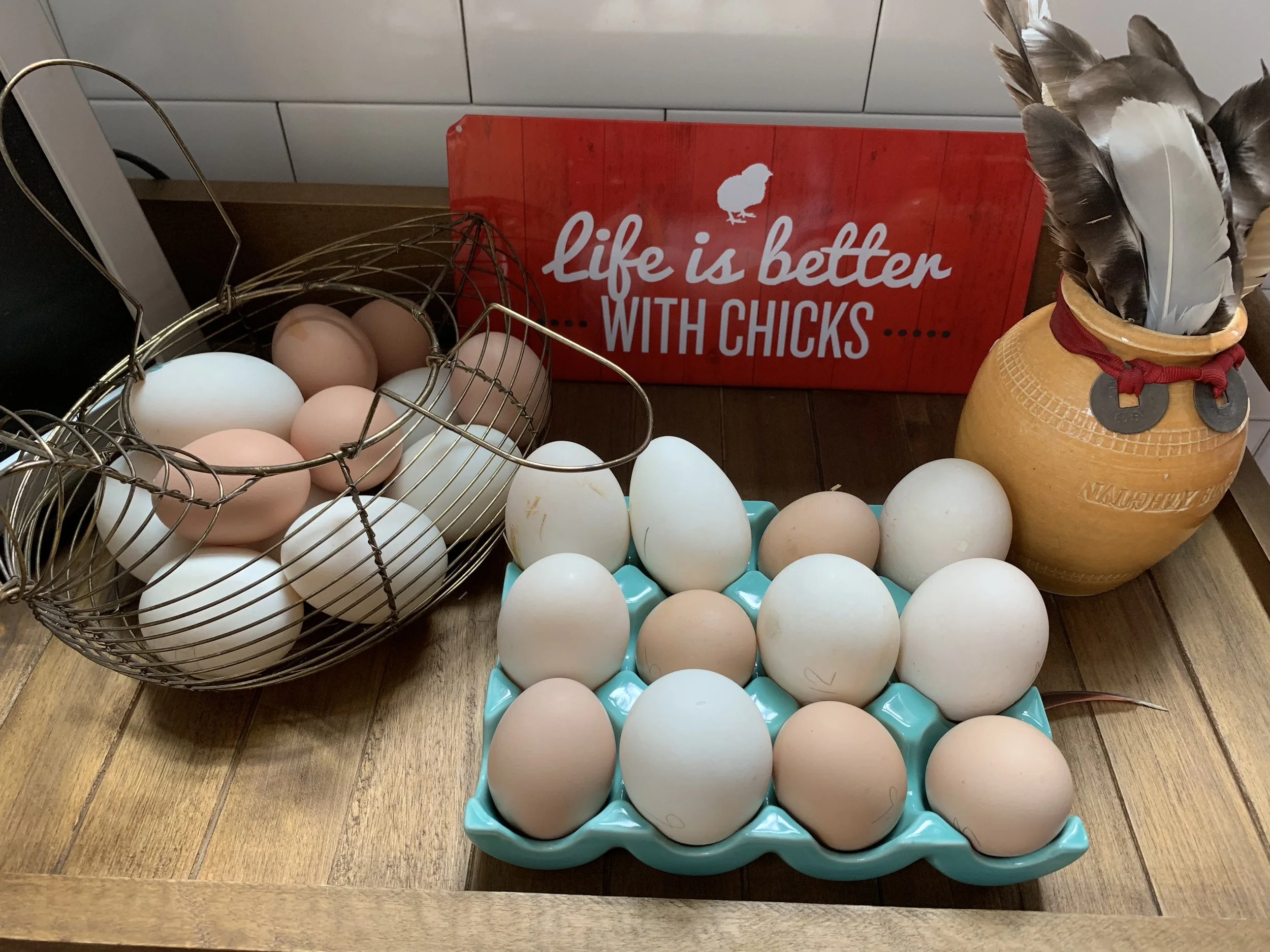 A wooden table displaying a basket of brown and white eggs, a tray of white eggs, a red sign reading 'Life is better with chicks,' and a clay pot filled with black, white, and brown feathers.