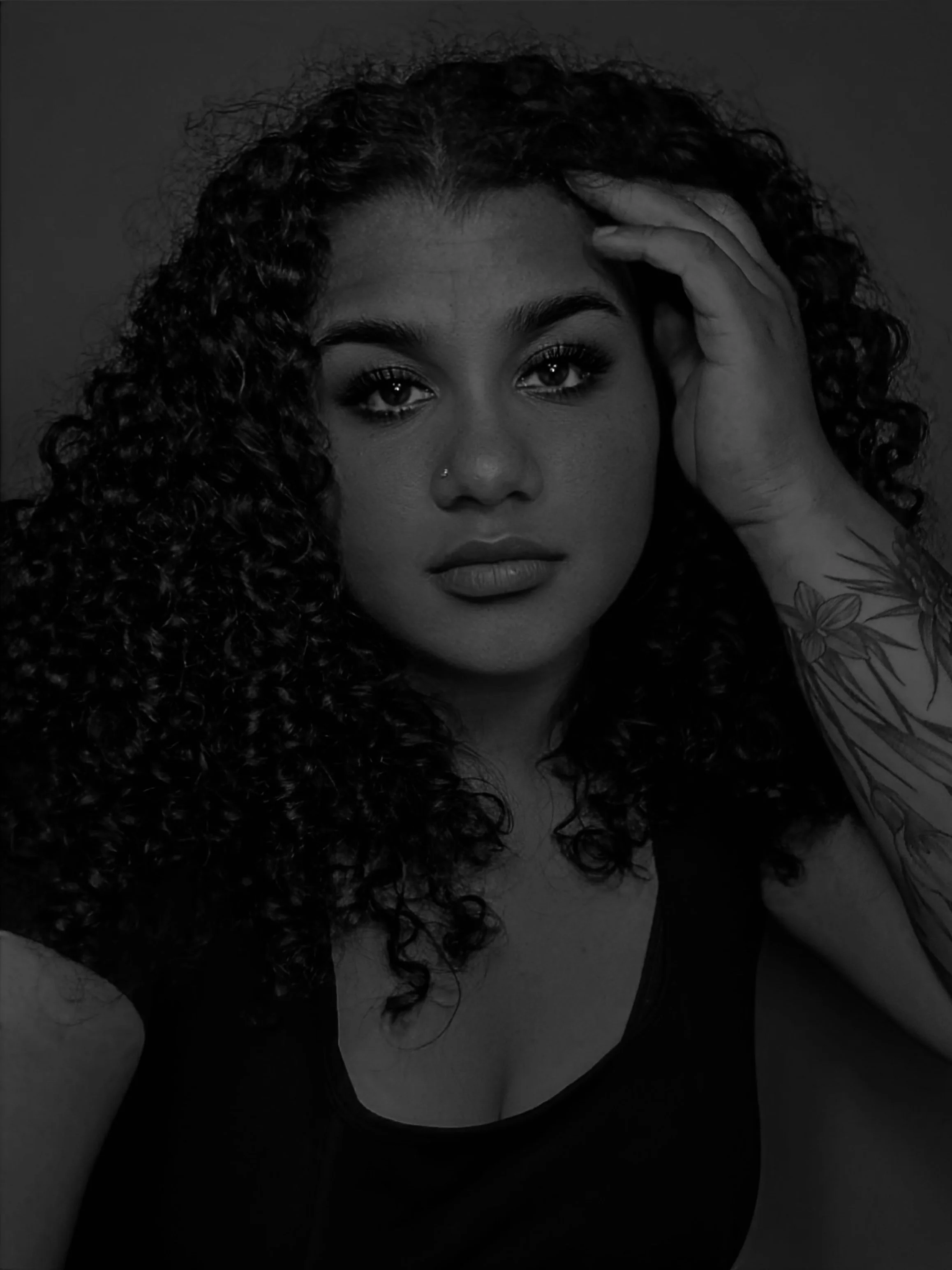 A woman with curly hair touching her forehead, looking directly at the camera, with tattoos on her arm, wearing a sleeveless top
