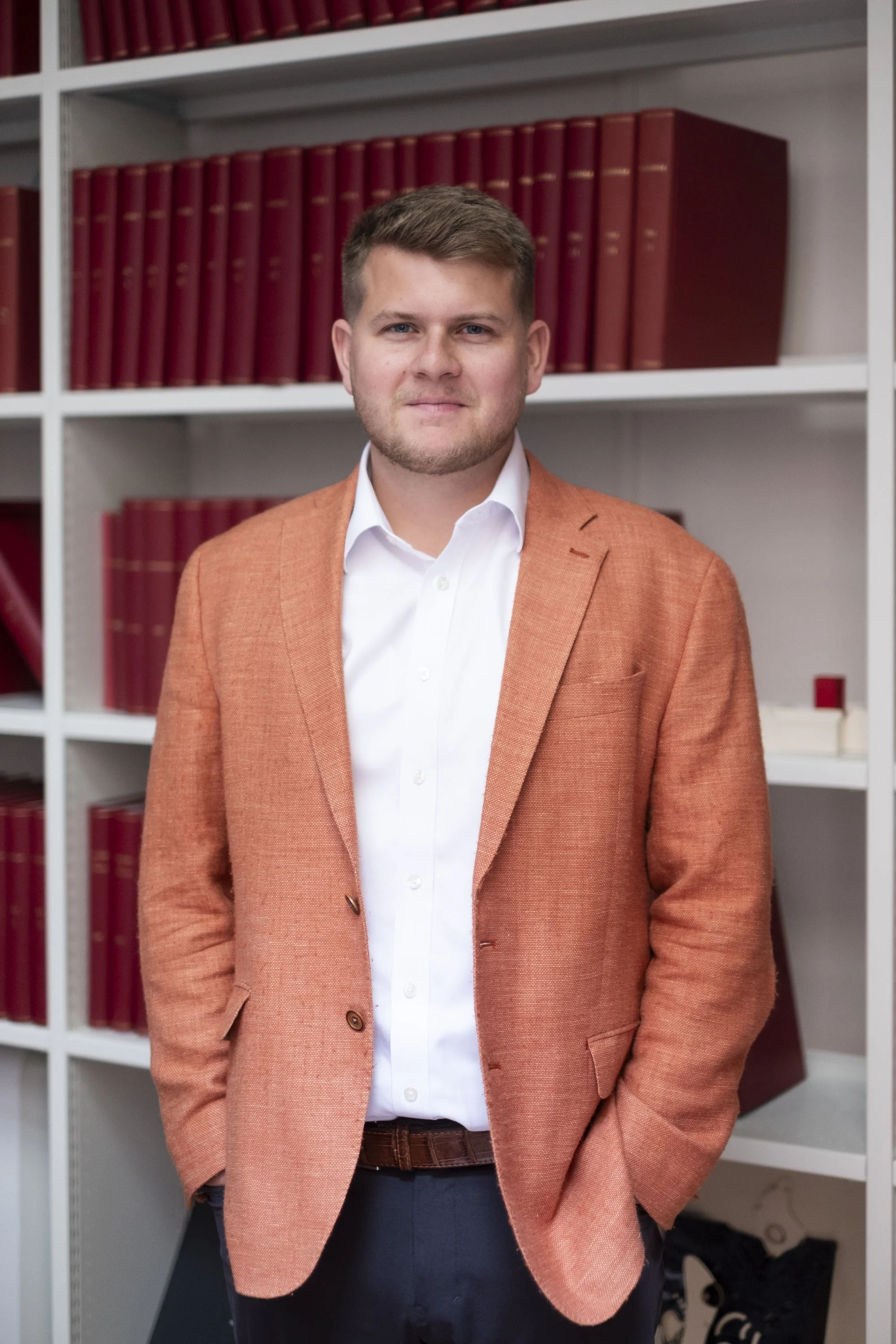 A man wearing a white shirt and orange blazer standing in front of a white bookshelf filled with red books.