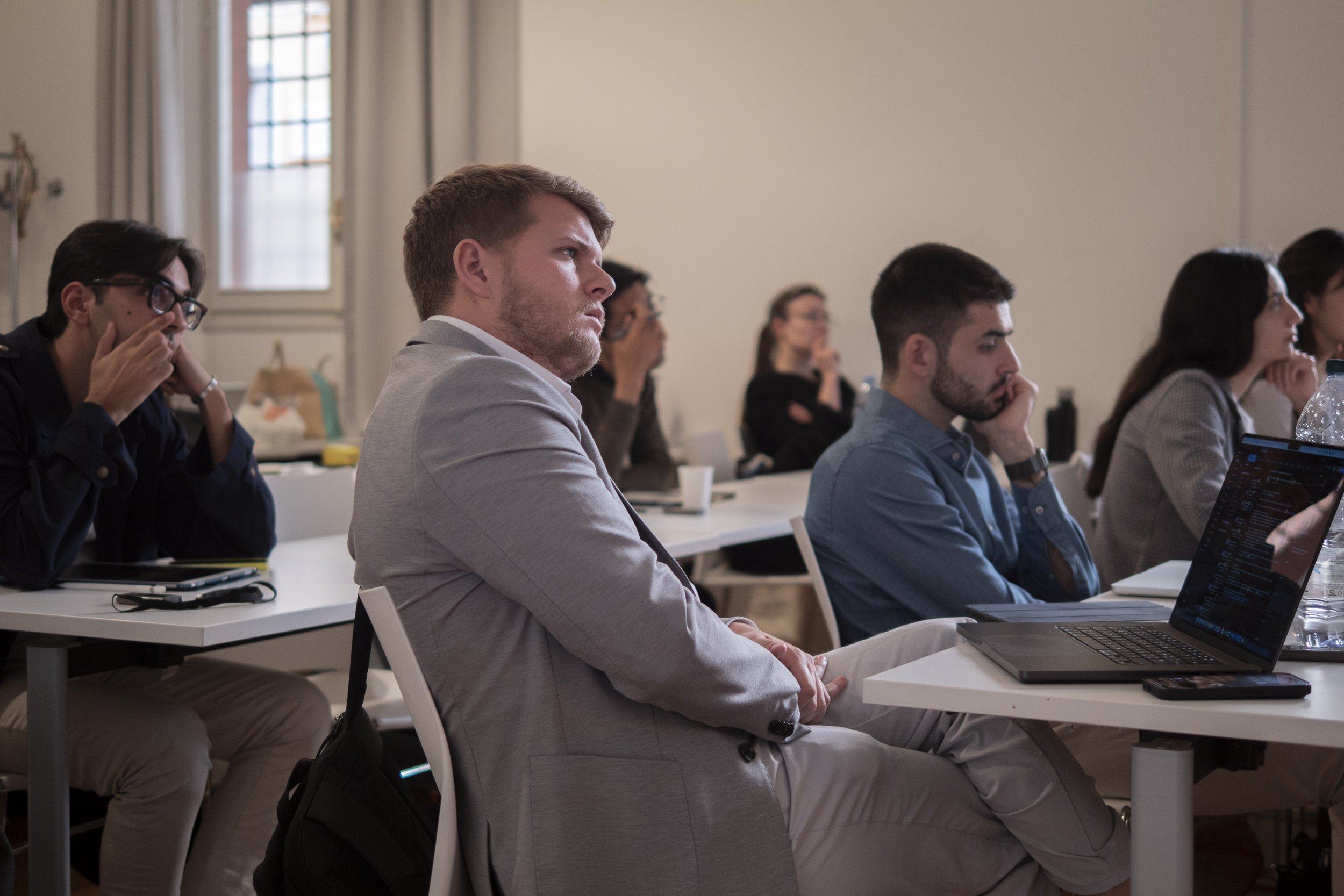 Group of people sitting at desks in a conference room, attending a presentation or lecture, some taking notes and others listening attentively.