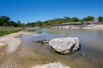 A shallow river with large rocks, surrounded by green trees and clear blue sky.