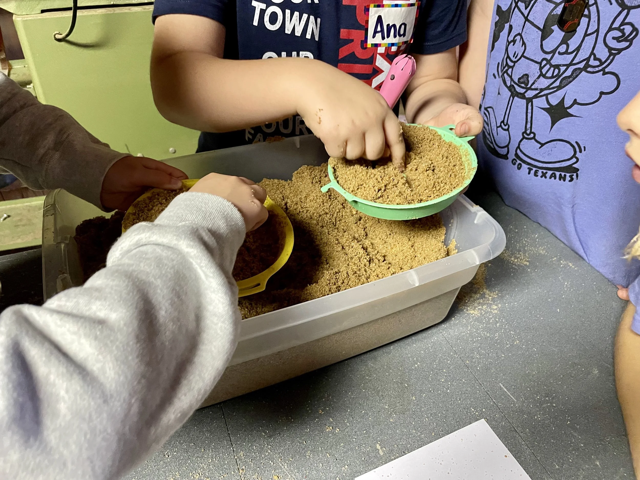 Children playing with kinetic sand in colorful containers at a table.
