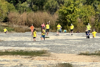 Group of children and adults wearing yellow shirts near a riverbank with trees in the background.
