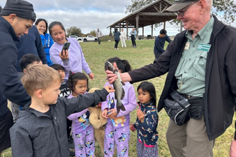A group of children and adults gathered outdoors under a pavilion, with some children receiving a small prize or gift from an older man.