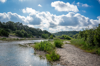 A peaceful river scene with a sandy shore, green vegetation, and a partly cloudy sky.