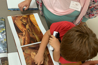 A young boy is examining dinosaur fossil images at a table, with an adult nearby.