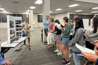 Group of people standing in a room, listening to a woman speak at a presentation or exhibit with display boards.