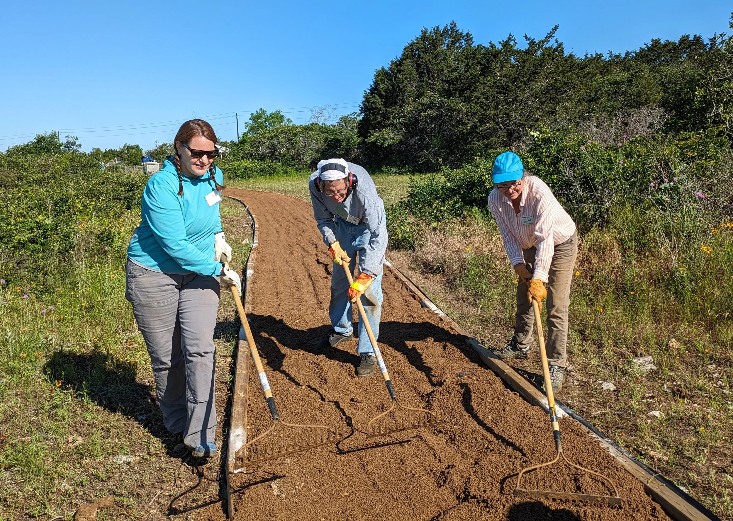Three people working on a dirt trail outdoors, leveling soil with rakes, surrounded by greenery and a clear blue sky.