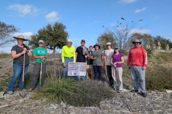 Group of people outdoors holding gardening tools, standing on a landscaped area with trees and a clear blue sky in the background.