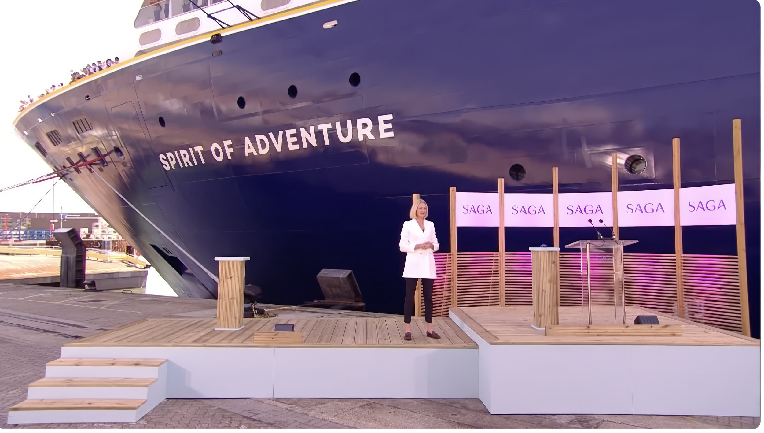 A woman stands on a wooden stage at a docked ship named 'Spirit of Adventure' with a backdrop of a cityscape. The stage has a podium with a microphone and banners with the word 'SAGA'.