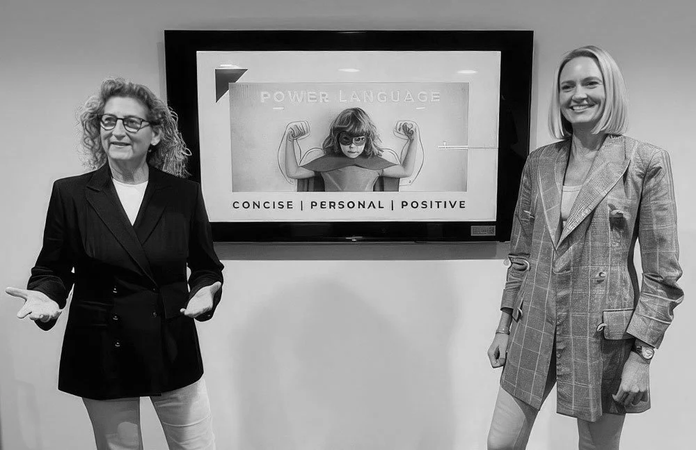 Two women standing on either side of a wall-mounted digital display, which shows a girl with glasses and a cape, flexing her arms with the text 'Power Language' above and 'Concise | Personal | Positive' below.