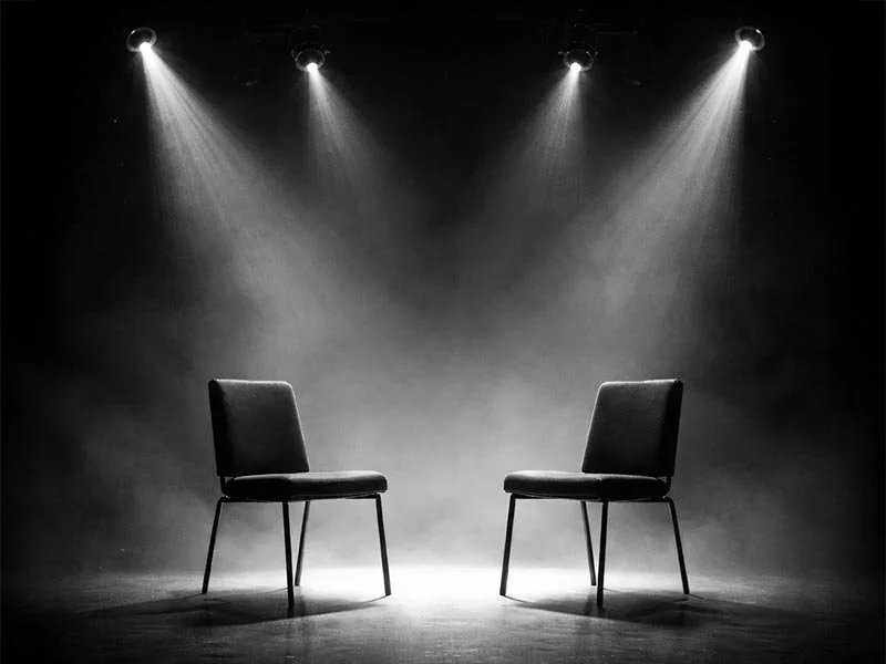 Two chairs facing each other on a stage with spotlights shining down, creating a dramatic and minimalistic setting.