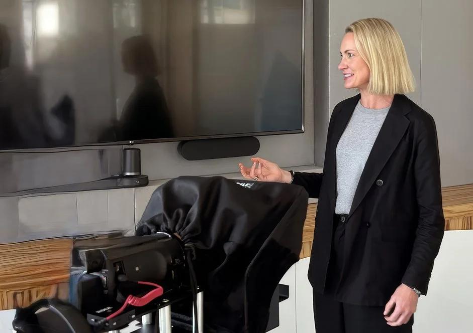 A woman with blonde hair in a black blazer and gray shirt, smiling and gesturing with her hand, standing next to a large flat-screen TV in a modern room.