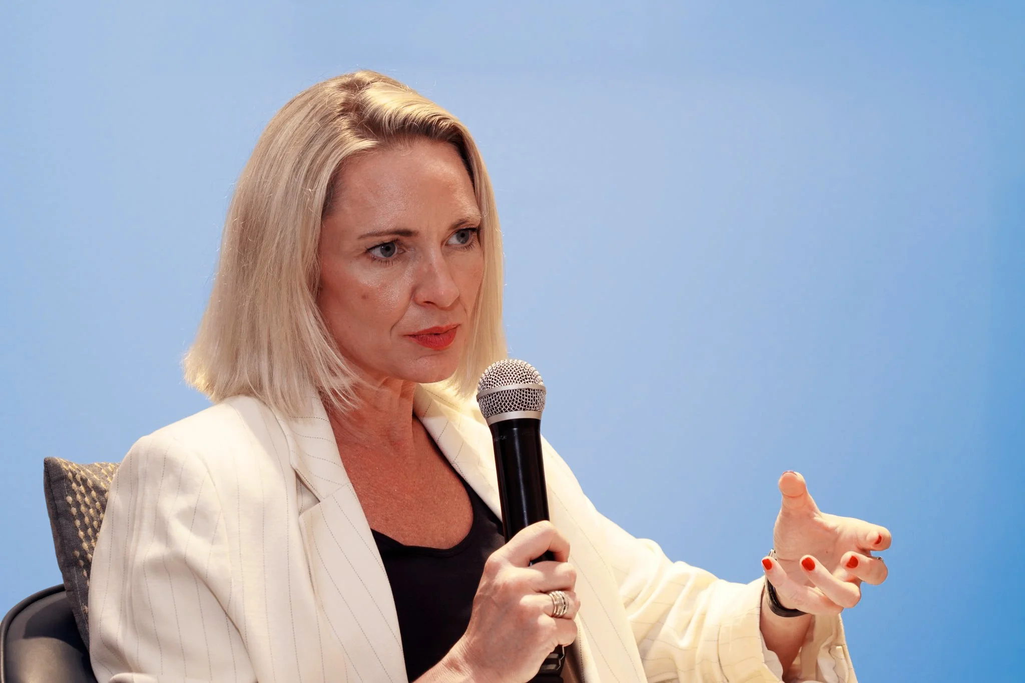 Heidi Mallace holding a microphone and speaking while gesturing with her right hand, against a plain light blue background.