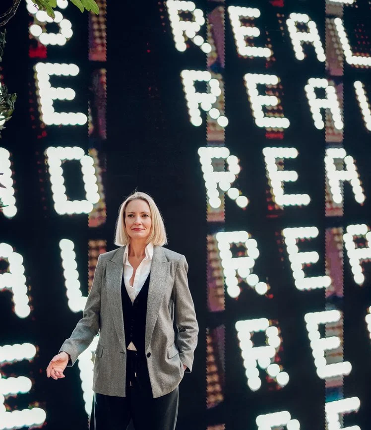A woman with blonde hair wearing a gray blazer and black pants, standing in front of a large illuminated sign with the words "Reel," "Real," and "Rebel".
