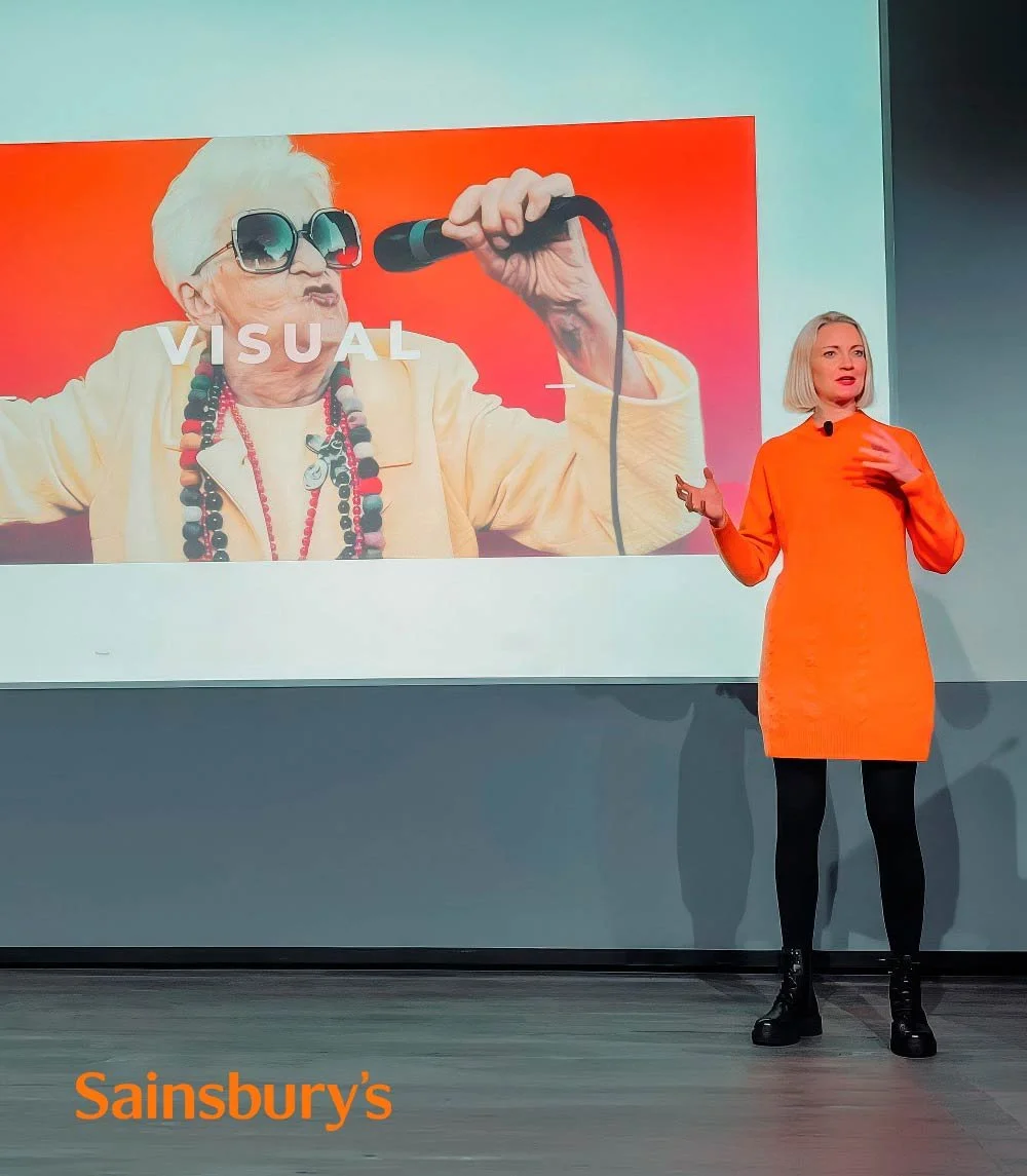 Heidi Mallace giving a presentation on stage with a large screen behind her displaying an elderly woman wearing sunglasses, holding a microphone, with the word 'VISUAL' over her. The Sainsbury's logo appears at the bottom left corner.