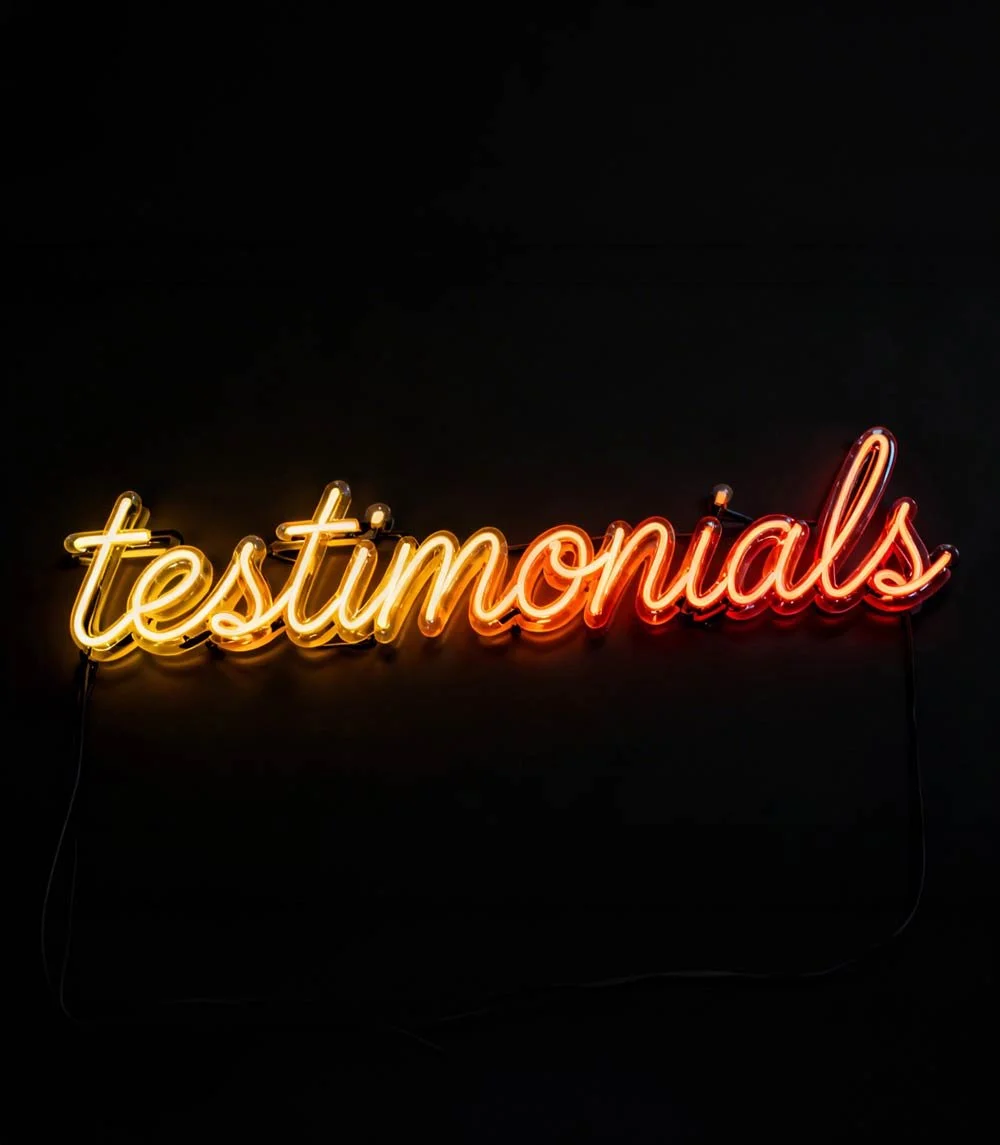Neon sign that reads 'testimonials' in yellow and red colors, mounted on a black wall.